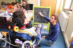 Ein Junge im Rollstuhl löst mit Hilfe eines Mitarbeiters Matheaufgaben auf einer Tafel.